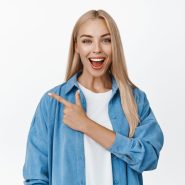 Amazed blond girl points finger left and looks impressed, shows advertisement, stands against white background Amazed blond girl points finger left and looks impressed, shows advertisement, stands against white background.